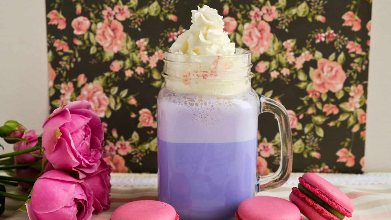 A clear glass mug filled with a purple-colored drink topped with whipped cream, is placed on a table. Surrounding the mug are four pink macarons and a small bouquet of pink roses. A black background with a pink floral pattern completes the scene.