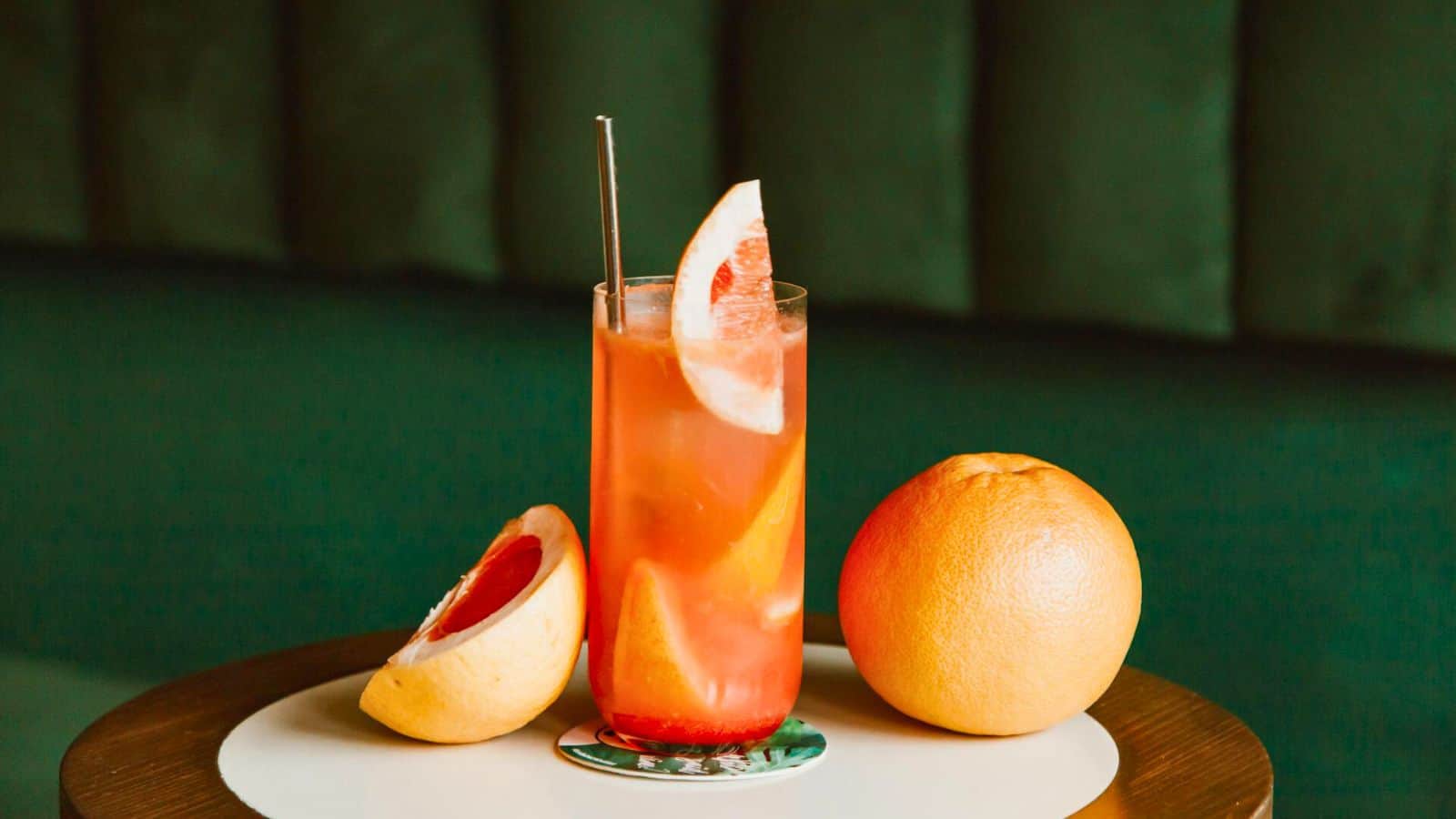 A tall glass of a pink-orange drink, garnished with slices of grapefruit and a metal straw, sits on a coaster on a round table. Next to the drink are a whole grapefruit and a half grapefruit. The background features a dark green upholstered surface.