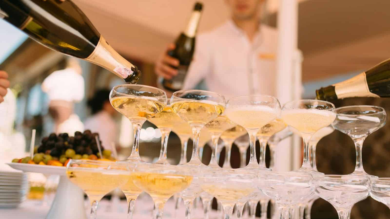 Several champagne glasses are stacked in a pyramid formation, with champagne being poured into the top glass. A person in a white shirt is holding a bottle of champagne. A plate with grapes and other fruits is placed next to the glasses.