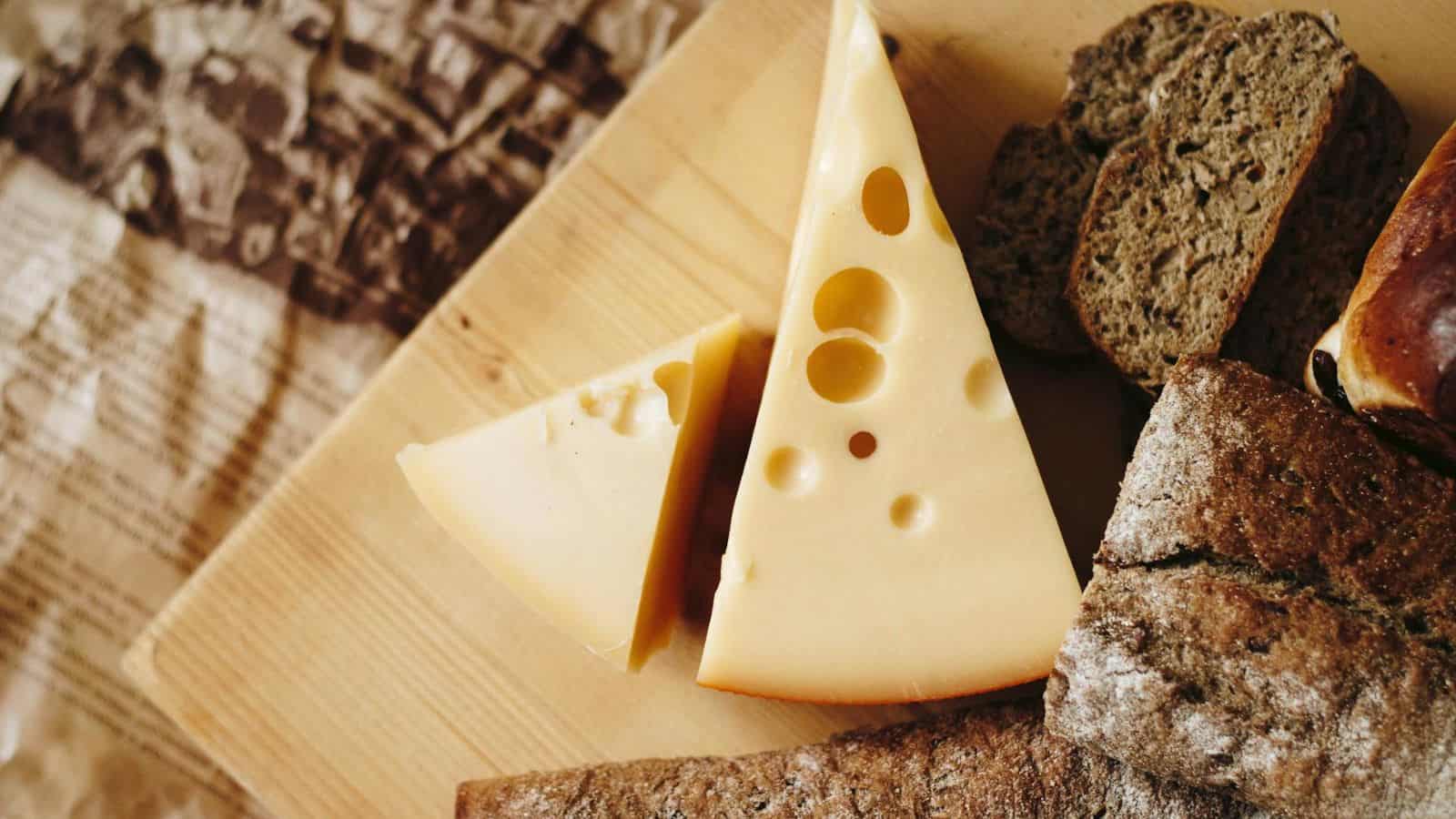 A cheese board with slices of Swiss cheese, featuring characteristic large holes, is accompanied by several pieces of brown bread. The board is placed on a light wooden surface with a textured fabric in the background.