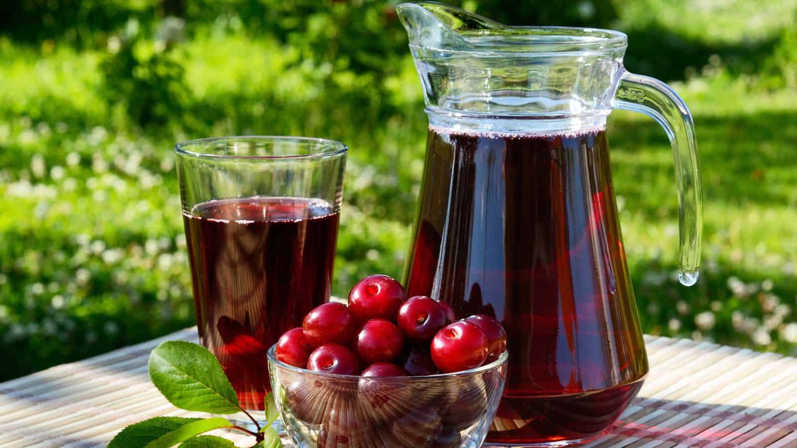 A clear glass pitcher and a glass of dark red juice sit on a light-colored, striped surface outdoors. A small, clear bowl of red cherries with green leaves rests in front of the pitcher and glass.