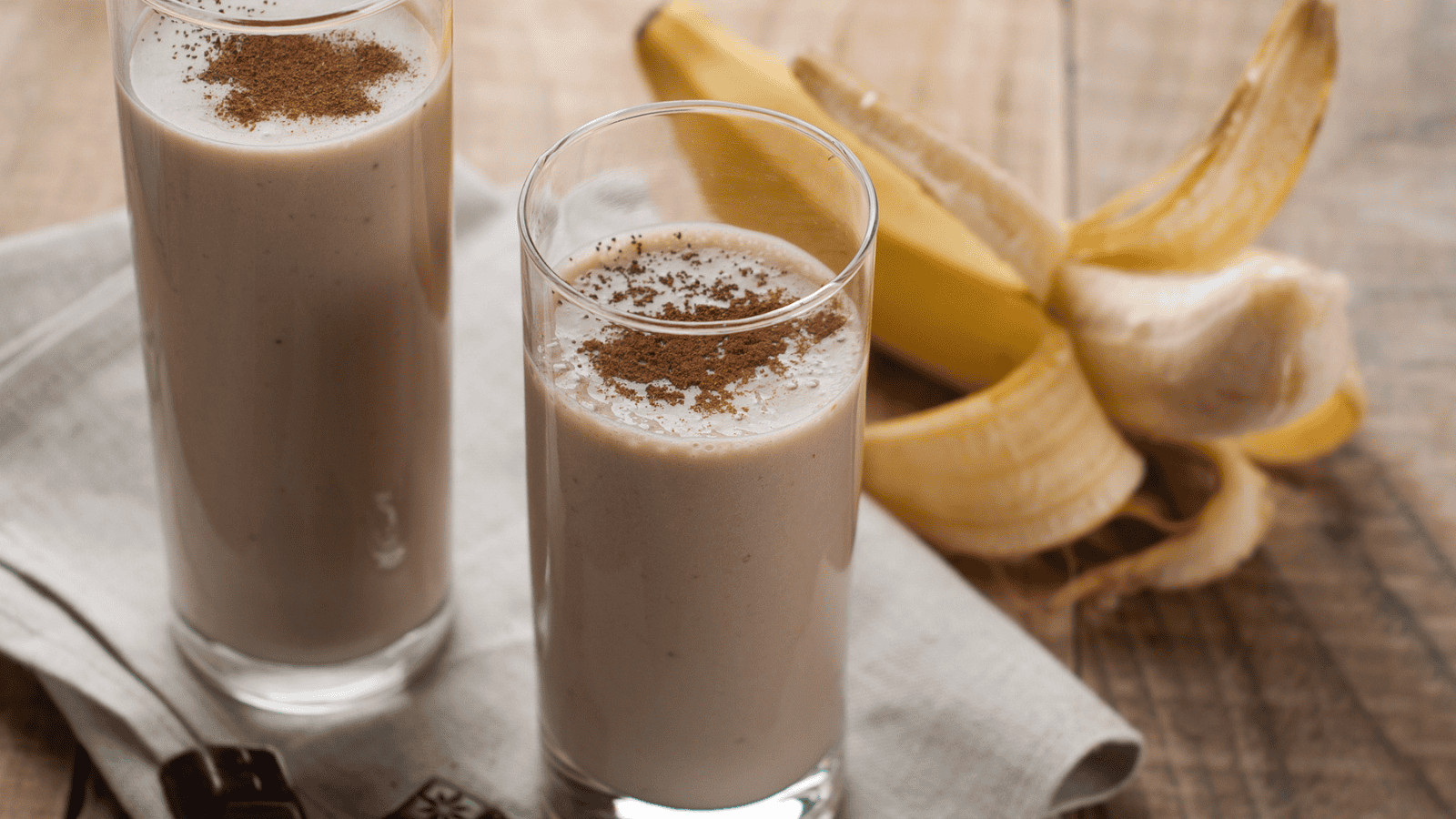 Two glasses of superfood banana smoothie topped with cinnamon sit on a wooden table. A peeled banana is nestled next to the glasses, with a napkin neatly placed underneath.