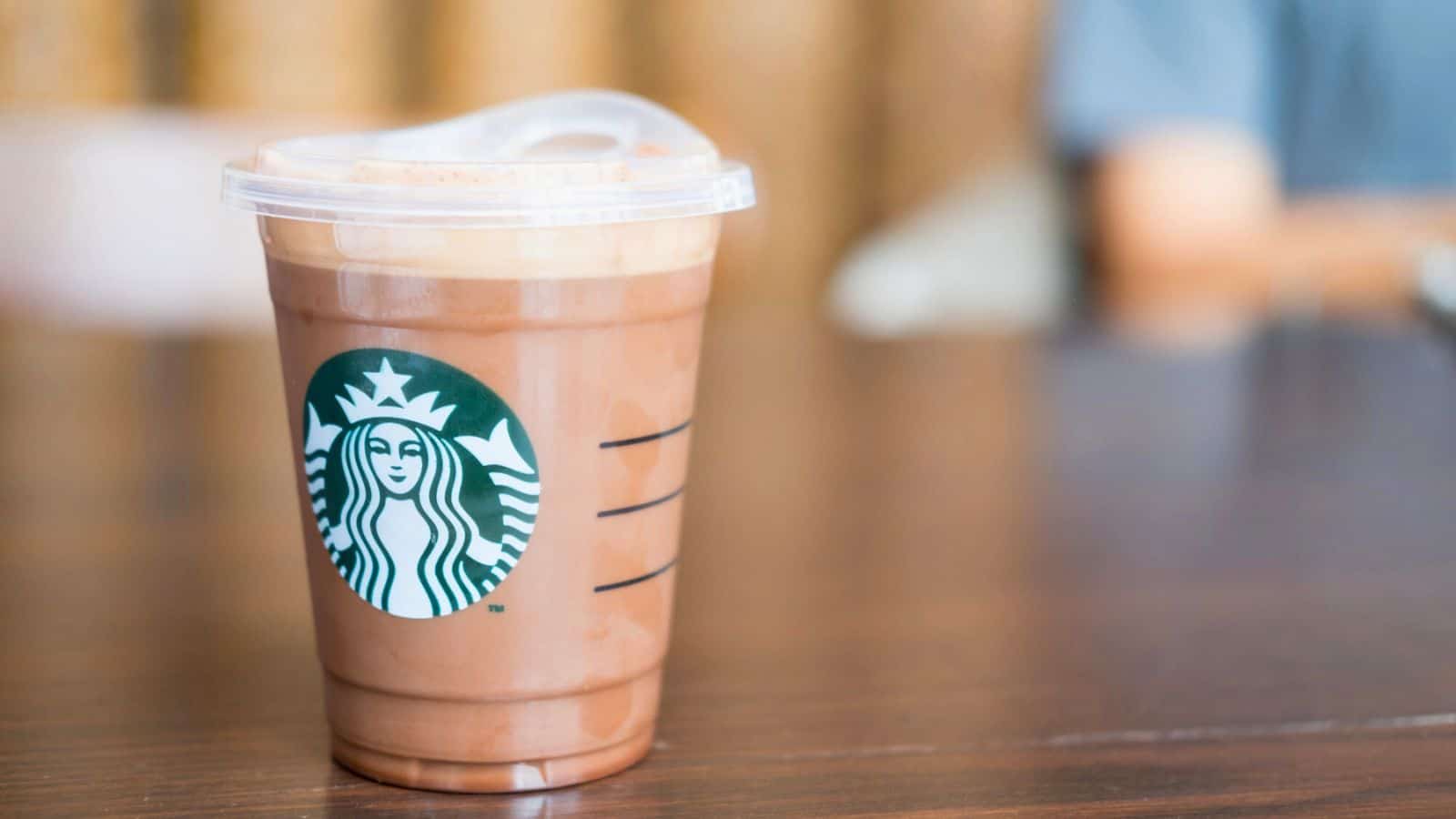 A Starbucks plastic cup filled with a chocolate beverage sits on a wooden table. The Starbucks logo is visible on the cup, and the background is blurred, showing a person sitting in the distance.