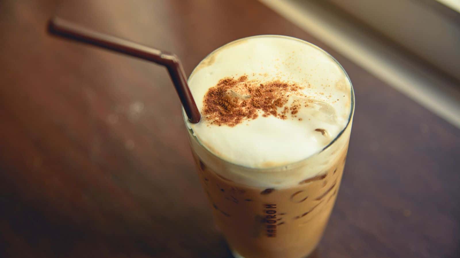 A glass of iced coffee topped with frothy milk and a sprinkle of cinnamon sits on a wooden table. A brown straw is inserted into the drink. Light streams in from a window, creating a bright ambiance. 