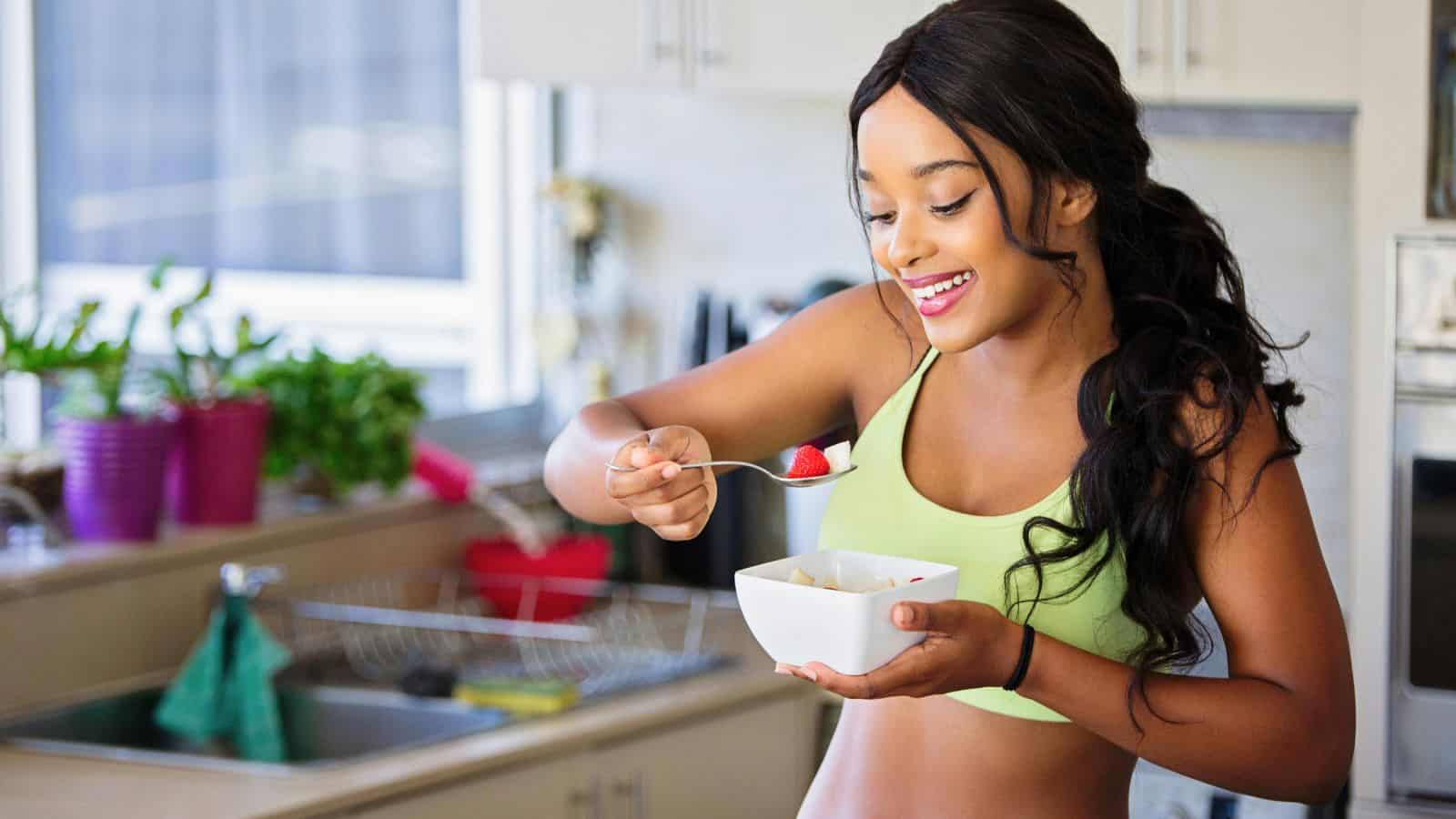A woman wearing a green sports bra smiles while holding a bowl of fruit in a kitchen. One hand holds a spoon with a piece of fruit, and the kitchen counter in the background displays plants and various kitchen items.