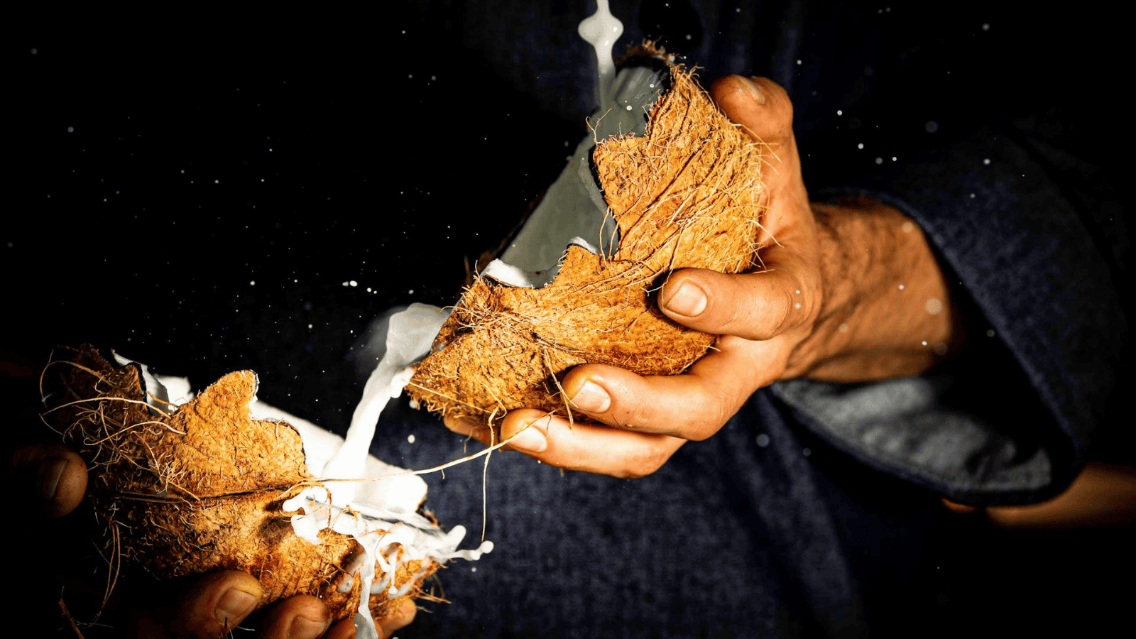 A person effortlessly splits open a coconut with their hands, releasing a splash of coconut water. The fibers and texture are vividly highlighted against the dark background, evoking the essence of plant-based dairy milk substitutes.