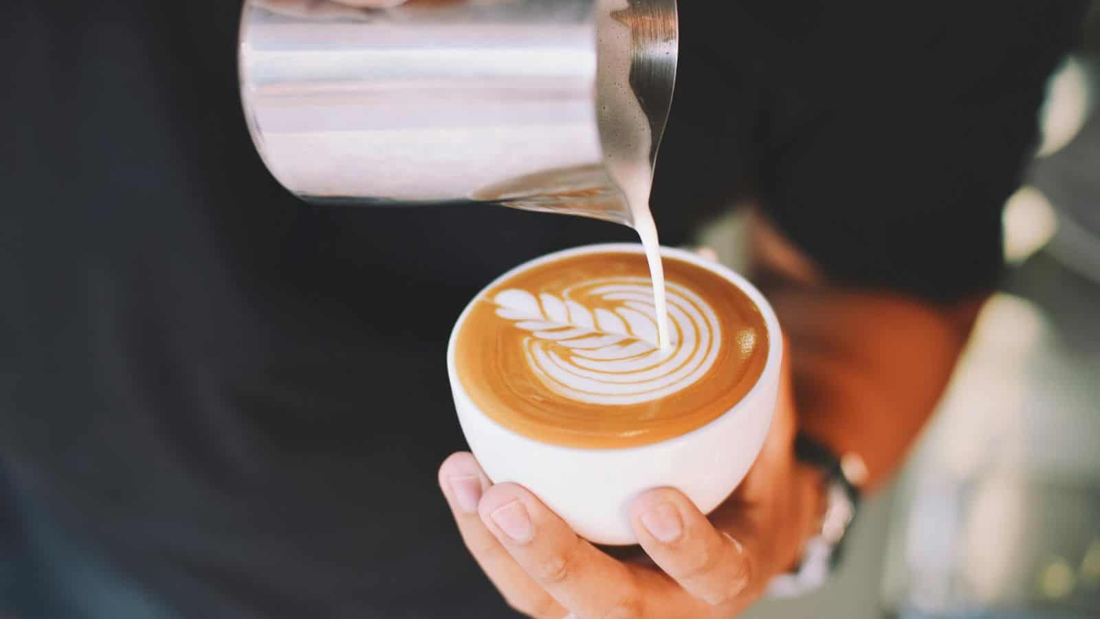 A barista is pouring steamed milk into a cup of coffee, creating a latte art pattern in the shape of a leaf. The barista's hand holds the cup steady while the other hand pours from a metal pitcher. 
