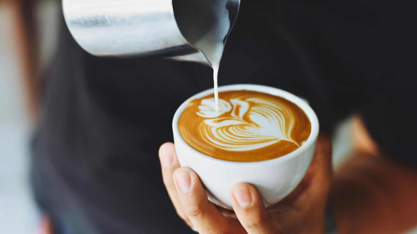 A person is pouring steamed milk from a metal pitcher into a cup of coffee, creating latte art in the shape of a flower. The person's hands are visible, and the background is blurred. The scene focuses on the process of making the latte art.