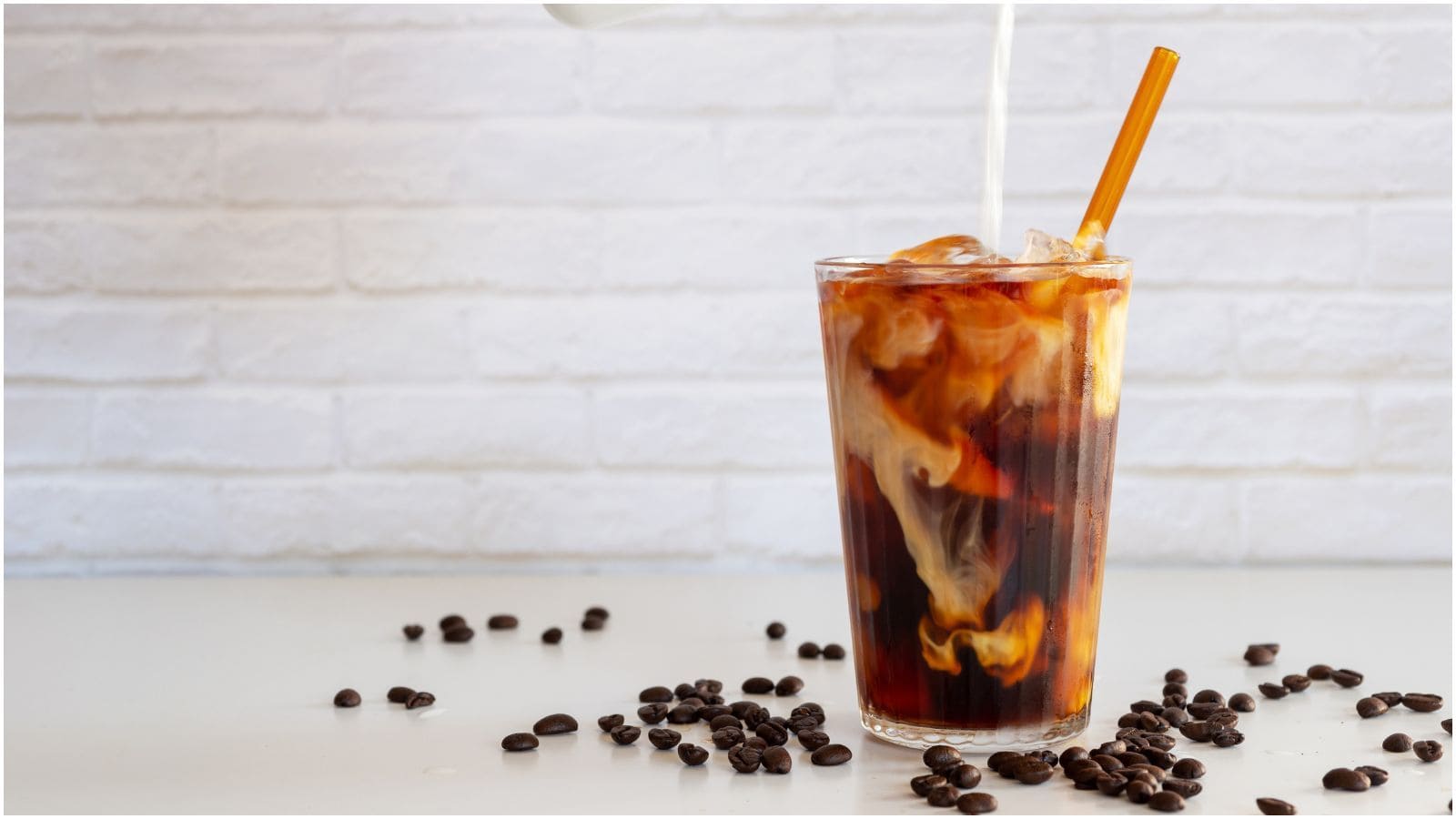 A glass of iced coffee is being poured with cream, creating swirls in the dark liquid. The drink has a straw, and coffee beans are scattered on the white surface in front of a white brick background.