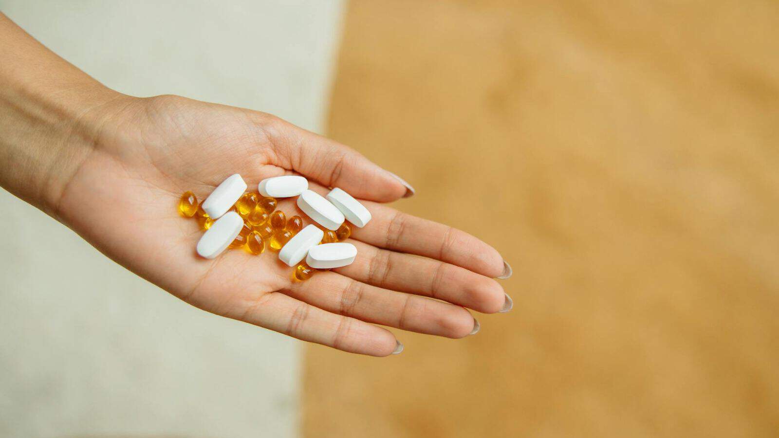 A hand is holding several white oval pills and yellow soft gel capsules against a blurred background.