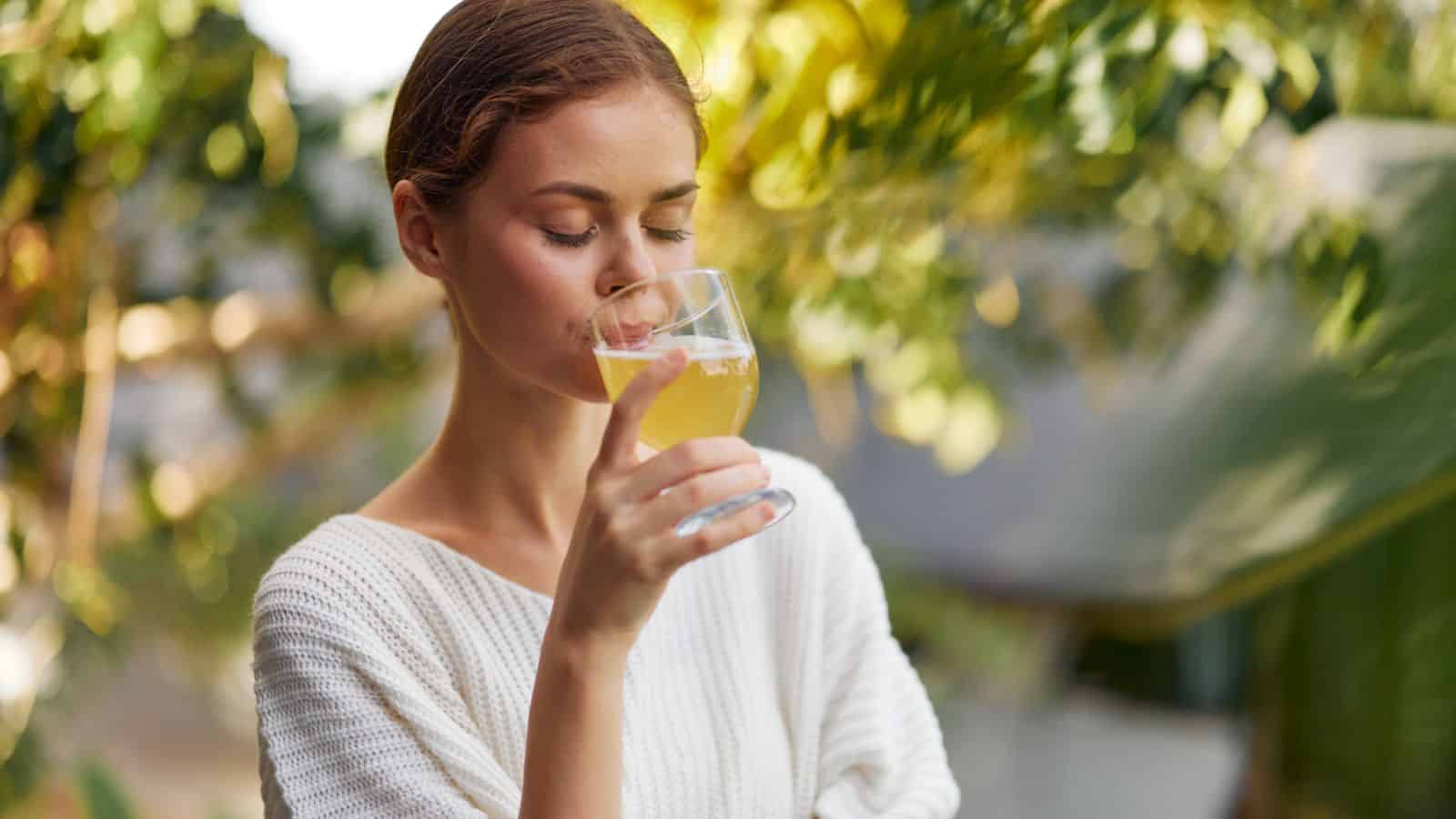 A woman sips kombucha from a clear glass. Her eyes are closed, and she appears to be enjoying the beverage. The background features blurry greenery, suggesting an outdoor setting.