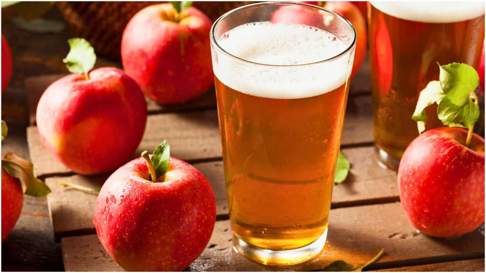 A glass of apple cider is placed on a wooden surface, surrounded by whole red apples. Another glass of cider is partially visible in the background. The cider has a frothy top layer, indicating it's freshly poured. The setting appears rustic and natural.