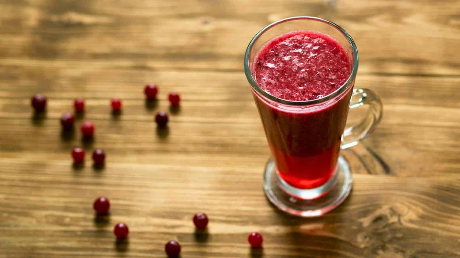 A tall glass filled with red cranberry juice is placed on a wooden surface. Several whole cranberries are scattered around the glass on the table. The juice appears smooth and glossy, with a rich, deep red color.