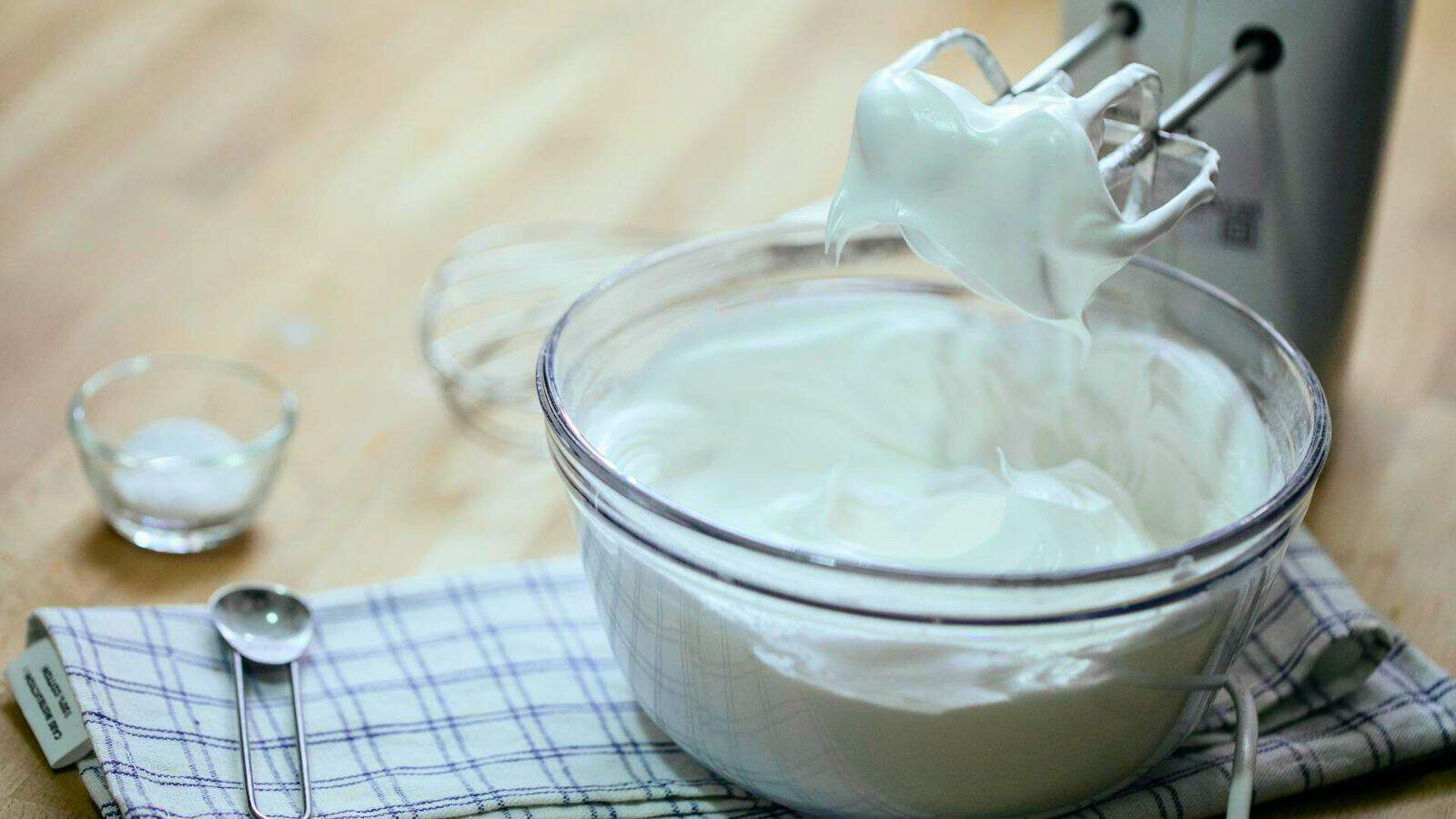 A glass bowl filled with whipped cream sits on a checkered cloth on a wooden surface. A hand mixer with beaters covered in whipped cream is positioned above the bowl. A small glass bowl and a metal teaspoon are nearby.