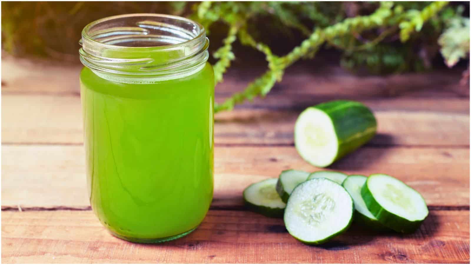 A glass jar filled with green juice is placed on a wooden surface. Next to it, there are several slices of cucumber and a partially sliced cucumber. Green foliage is blurred in the background.