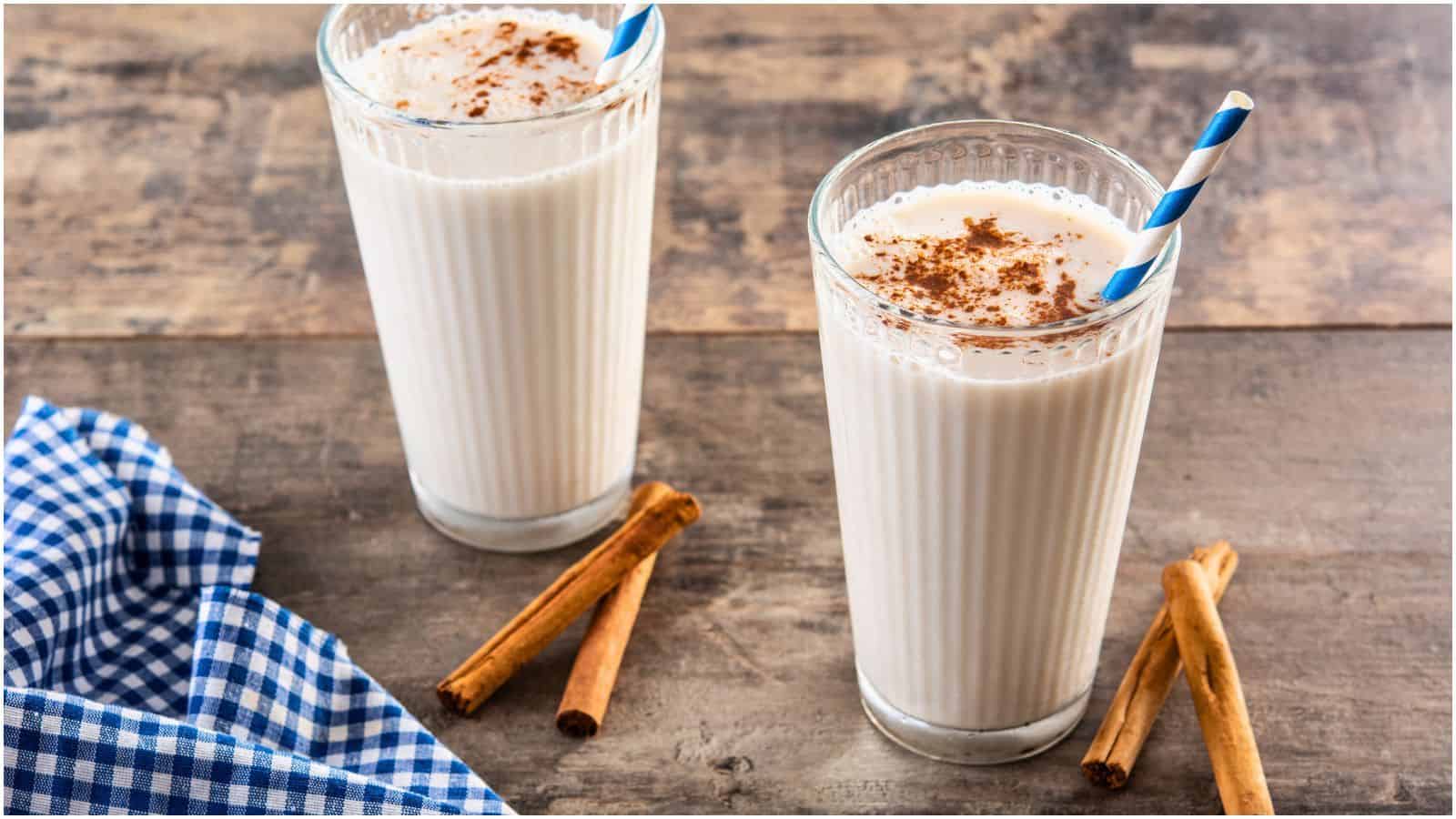 Two glasses of milk topped with cinnamon powder, each sporting a blue and white striped straw, rest on a wooden surface. Cinnamon sticks lie nearby, adding to the cozy vibe, while a blue and white checkered cloth is positioned to the left. 