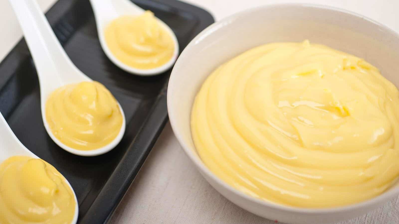 A bowl of smooth, creamy yellow custard is placed next to a black tray holding three white ceramic spoons, each with a small dollop of custard. The custard appears glossy and thick. The background is a neutral, light-colored surface.