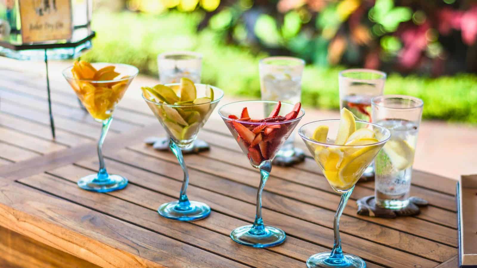 A wooden table holds four blue-stemmed martini glasses filled with sliced fruits, including strawberries, oranges, pears, and lemons. Behind them, three tall glasses of water with lemon slices are arranged in a row. The background is a blurred outdoor setting.