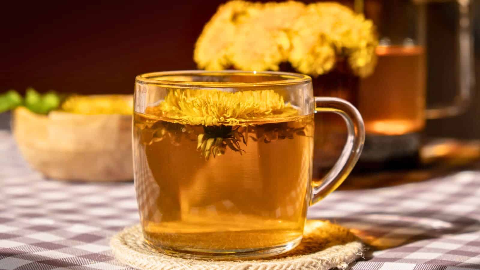 A clear glass mug filled with dandelion tea sits on a woven coaster. A yellow dandelion flower floats in the tea. In the background, there is a bowl with more flowers and another glass of this drink. The table is covered with a checkered tablecloth.