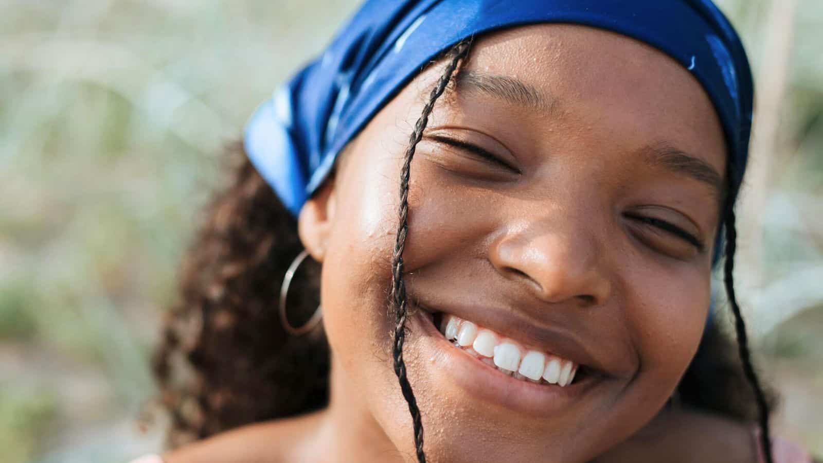 A person with braided hair and a blue headscarf smiles broadly. The background is blurred, suggesting an outdoor setting. The person is wearing hoop earrings.