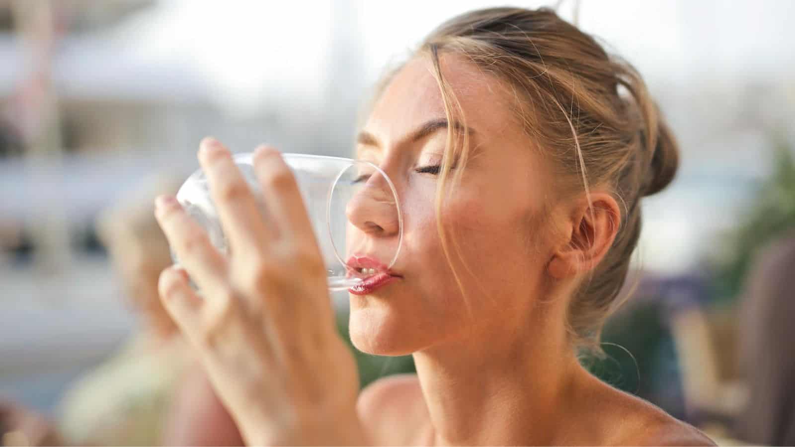 A woman with blonde hair is drinking from a clear glass, her eyes closed. She is sitting outdoors, and the background is softly blurred, suggesting a waterfront or marina setting.