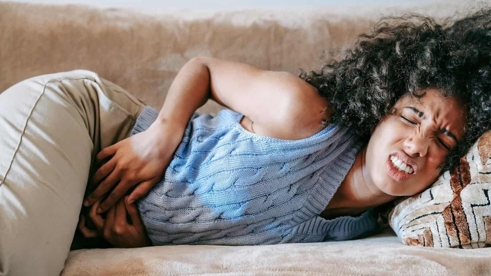 A person with curly hair wearing a light blue sweater and beige pants lies on a beige couch with a pained expression and hands clutched to their abdomen. 