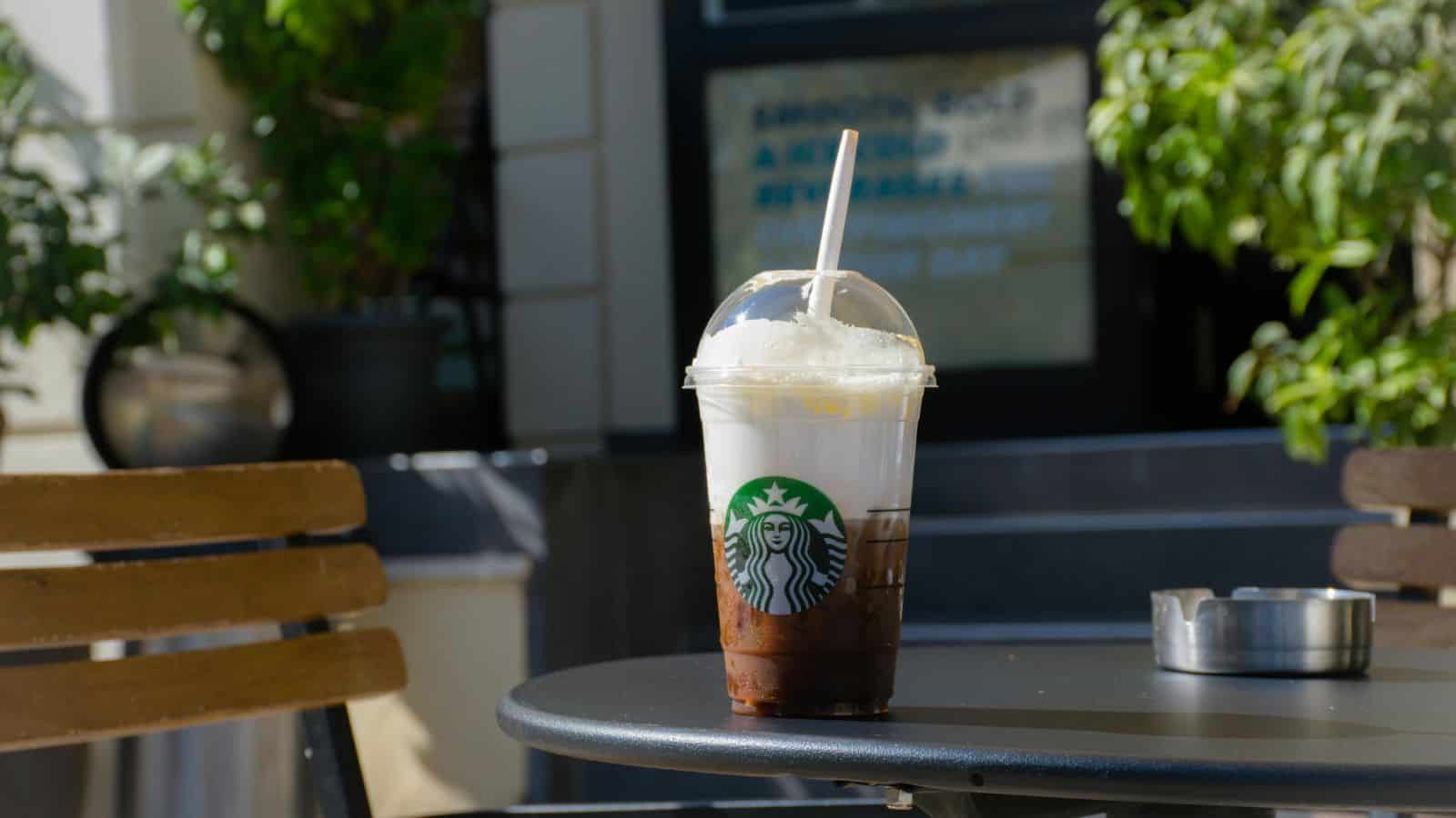 A plastic cup filled with an iced coffee drink is placed on a black outdoor table near a wooden chair. The cup, branded with the Starbucks logo and straw. The background includes a potted plant and a building wall with some indistinct signage.
