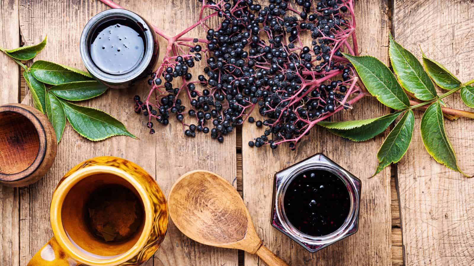 A wooden table with two jars of elderberry jam, fresh elderberries on their stems, a wooden spoon, a wooden mortar, and green leaves arranged around.