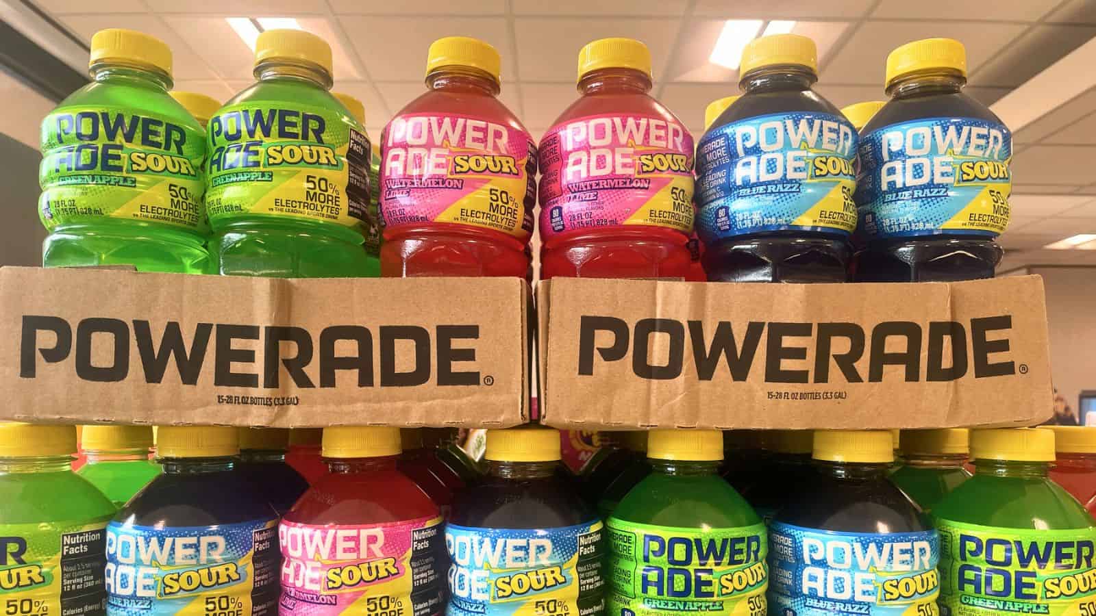 A display of Powerade Sour drinks in a store, featuring green, yellow, pink, blue, and purple bottles. The bottles are stacked in rows, with cardboard boxes labeled "Powerade" visible in the middle. 
