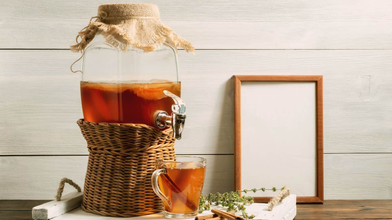 A glass beverage dispenser filled with kombucha sits on a wicker stand. 