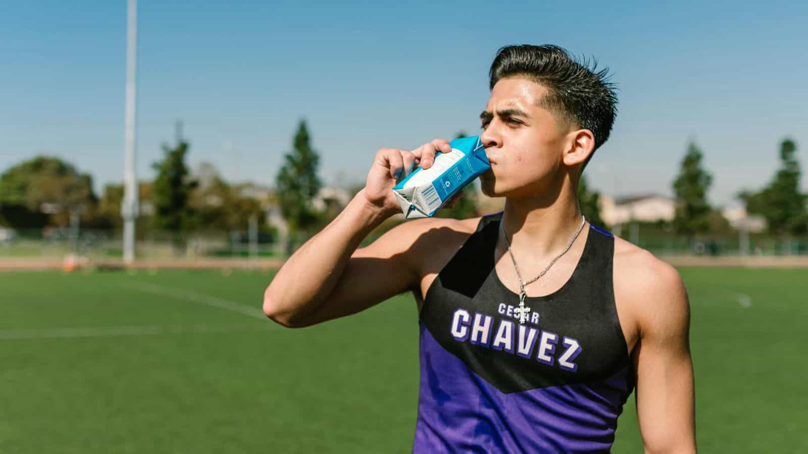 A person wearing a black and purple athletic top labeled "Chavez" stands on a field, drinking from a blue and white carton. The individual appears to be taking a break from physical activity, with trees and buildings visible in the background.