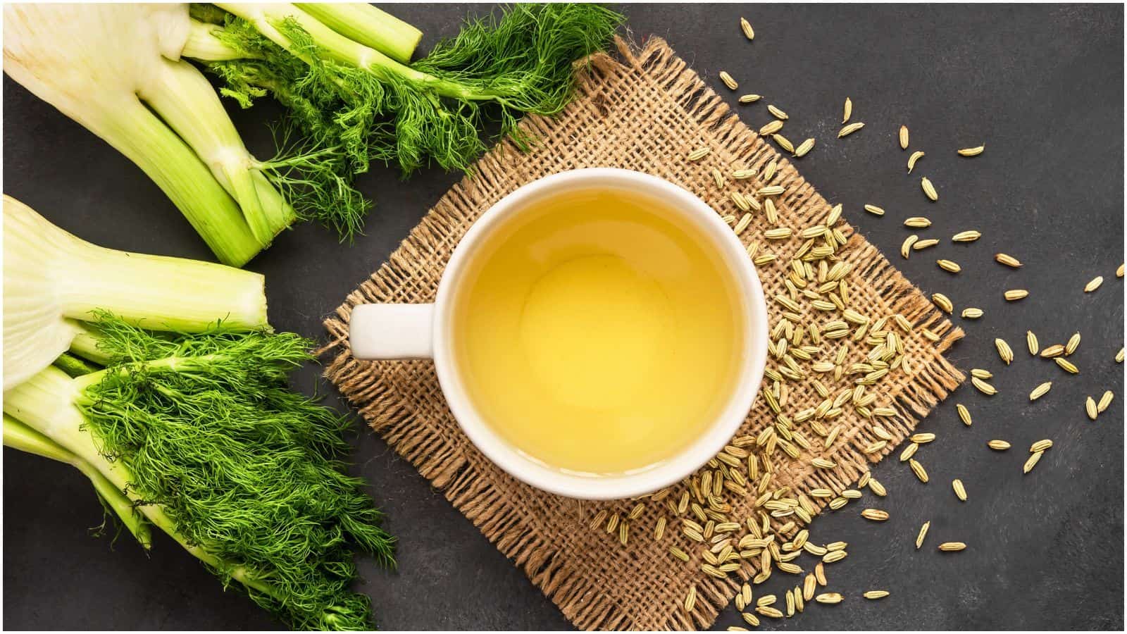 A white cup containing yellow liquid is placed on a woven mat on a dark surface. Surrounding the cup are fennel seeds and fresh fennel stems with green fronds.