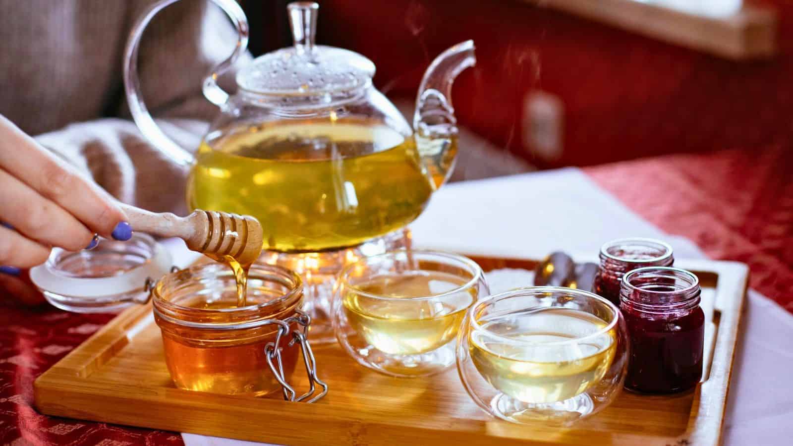 A person is dipping a honey dipper into a jar of honey next to a teapot and two teacups filled with tea. There are also two small jars of jam on the wooden tray, which is set on a red tablecloth.
