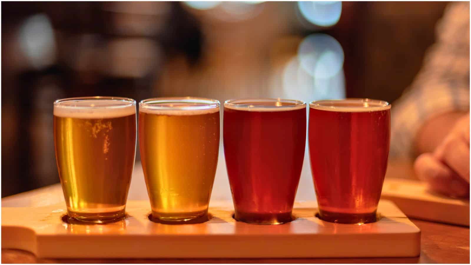 A close-up of a wooden tray holding four small glasses of non-alcoholic beer in varying shades of amber, from light to dark. The background is softly blurred, and a person's arm is partially visible to the right.