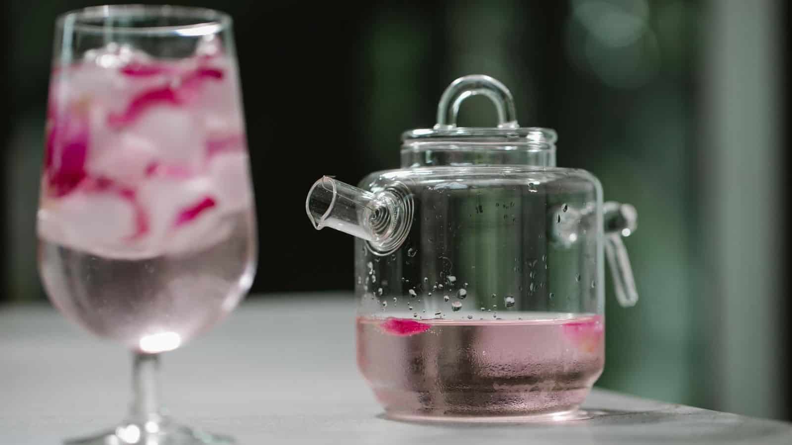 A clear glass teapot with a pink beverage and a glass with ice and pink flowers inside it are placed on a white surface. The background is blurred with a green nature setting, likely trees or plants.