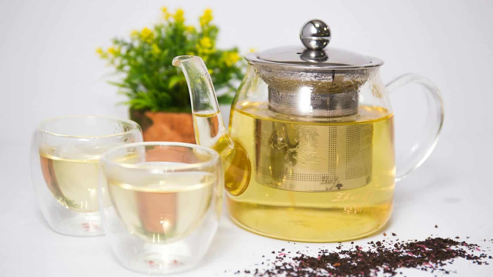 A glass teapot filled with tea and a metal infuser sits next to two glass teacups containing tea