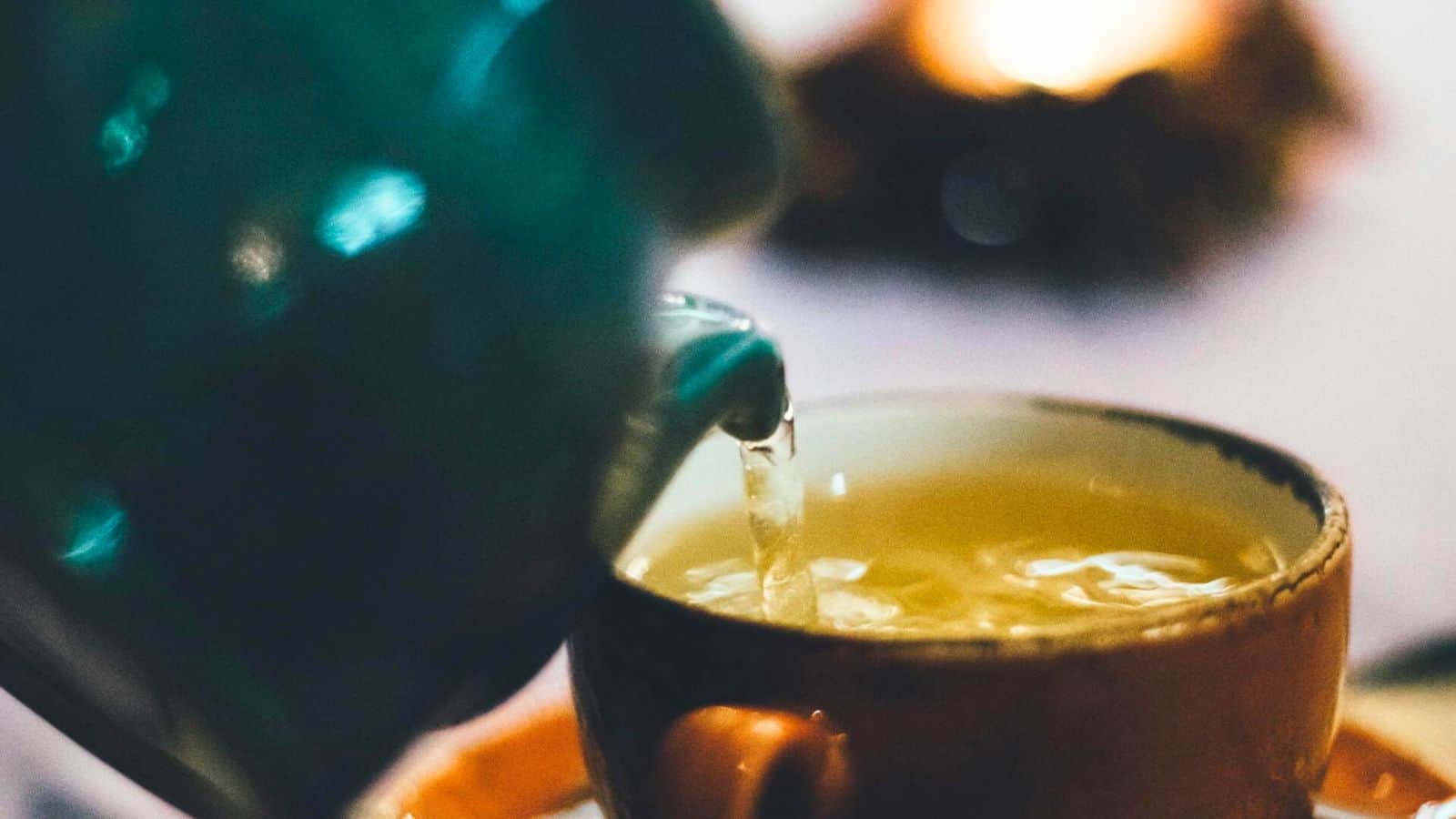 A close-up image of tea being poured from a blue ceramic teapot into a brown ceramic teacup. The liquid is light yellow and the background is slightly blurred, with warm lighting.
