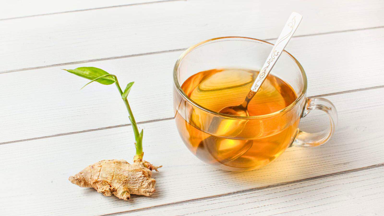 A glass cup of tea with a spoon sits on a white wooden surface. Next to the cup, there is a piece of ginger with a green sprout growing from it.