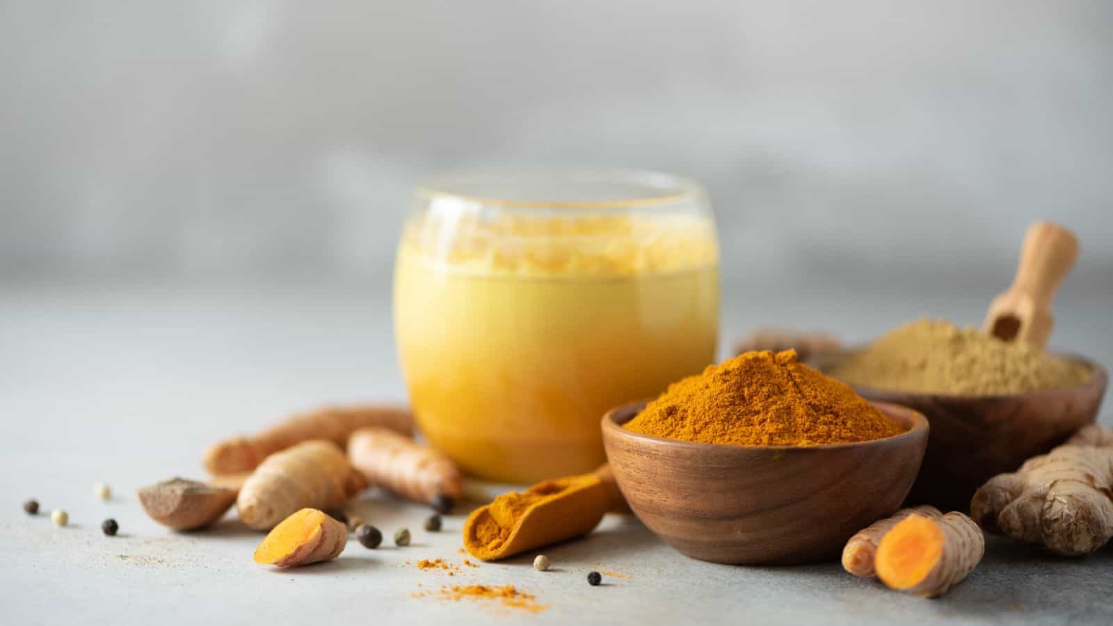 A soothing glass of turmeric milk surrounded by fresh turmeric roots, a wooden bowl brimming with turmeric powder, another bowl with a different spice powder, and scattered peppercorns on a light background.
