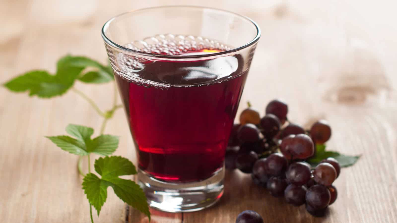 A glass of red grape juice sits on a wooden surface, next to a small bunch of dark grapes and green vine leaves.