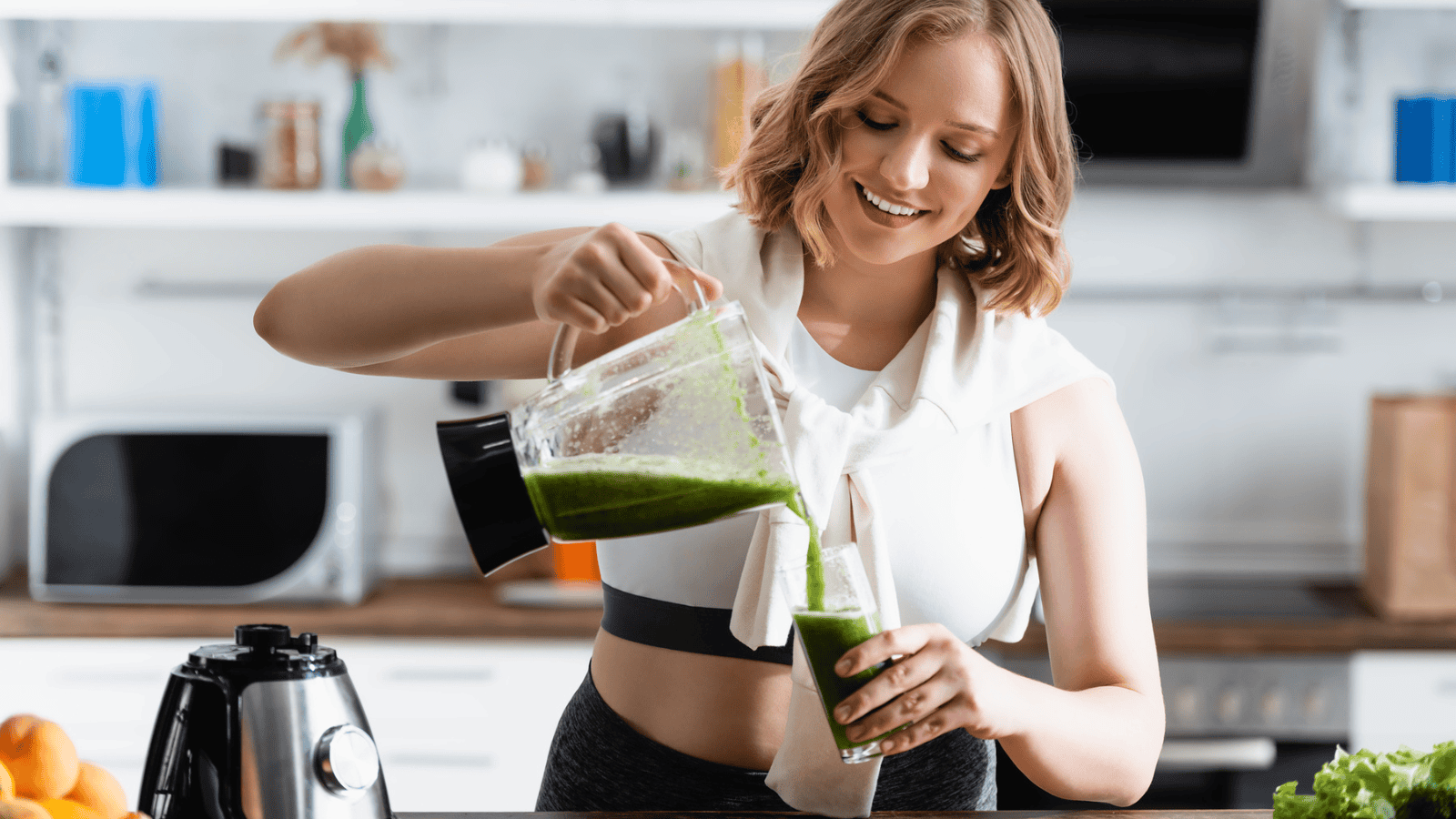 A woman in a kitchen pours a superfood smoothie from a blender into a glass. The kitchen counter, adorned with fruits and the blender base, complements her lively scene. She wears a white top and black athletic pants, smiling at her task.