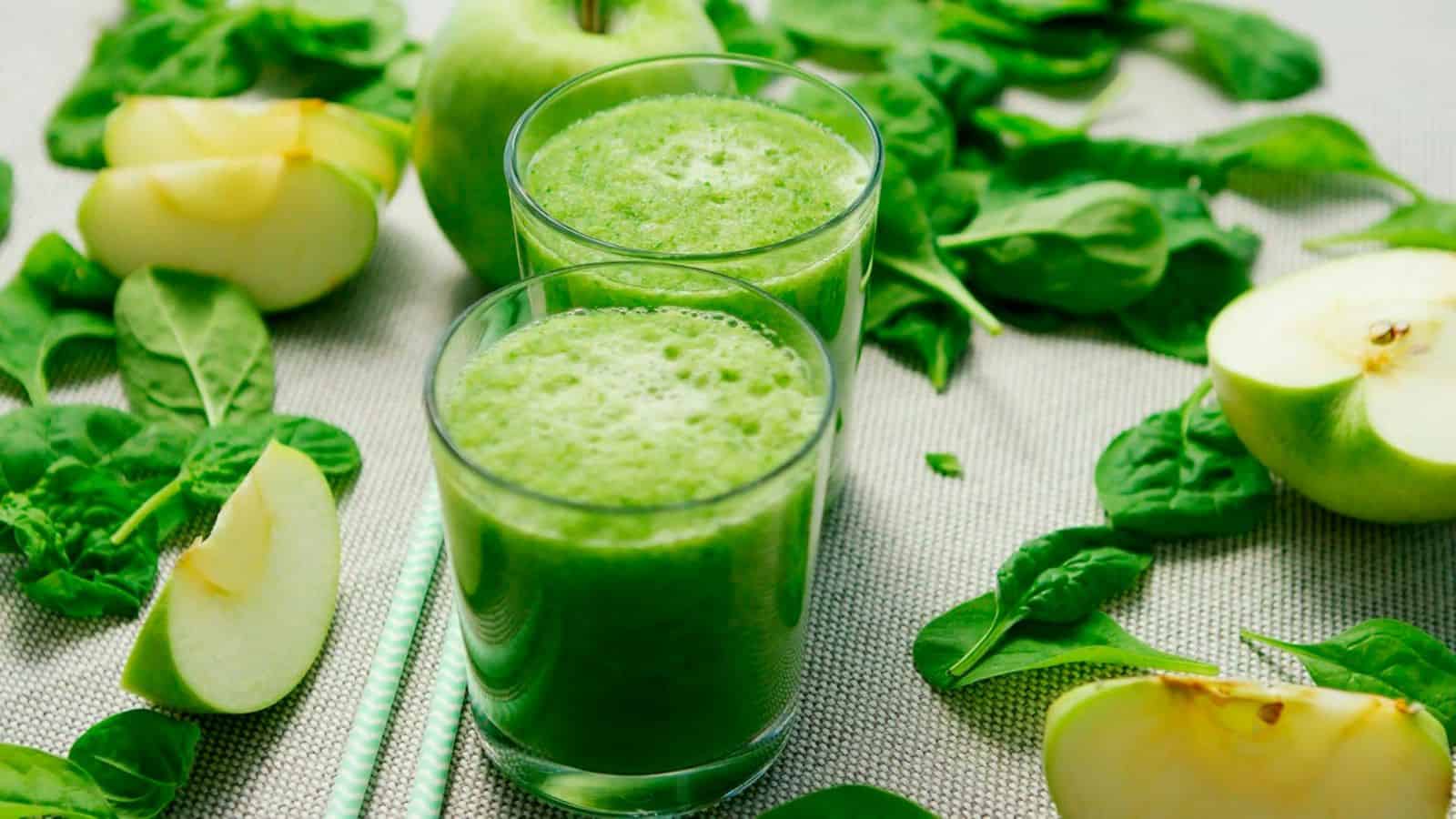 Two glasses of green smoothie are placed on a table surrounded by fresh spinach leaves, whole green apples, and cut apple slices. Two green-striped straws lie on the table beside the glasses.