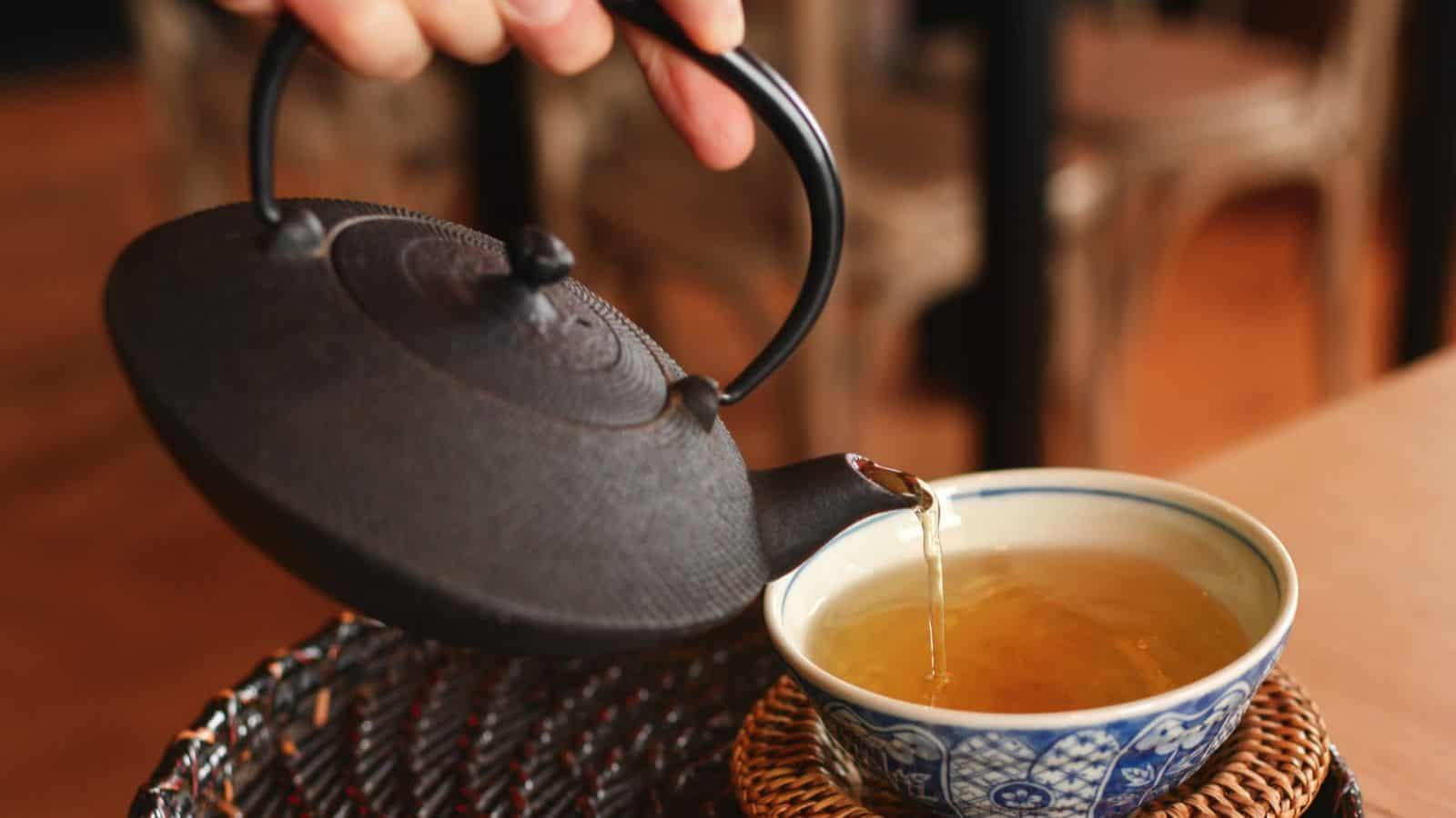 A hand pouring tea from a black cast iron teapot into a patterned ceramic cup placed on a woven coaster on a wooden table.