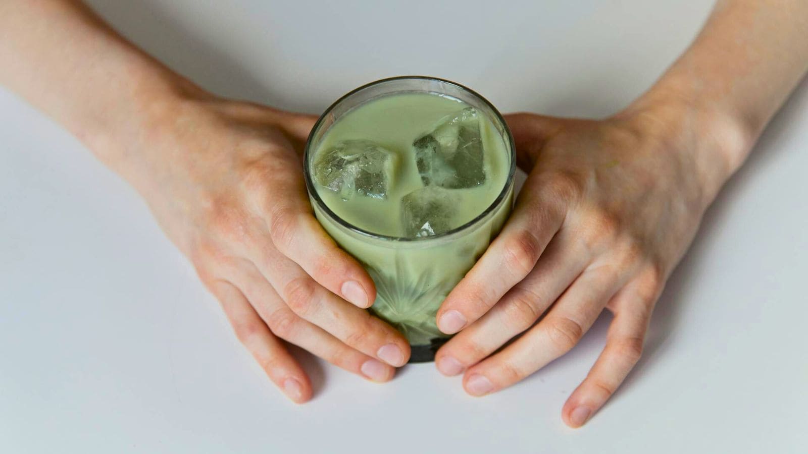A person is holding a glass filled with a light green beverage and ice cubes. The hands are gently wrapped around the glass, which is placed on a white surface. The drink appears to be a cold matcha latte or similar green liquid.