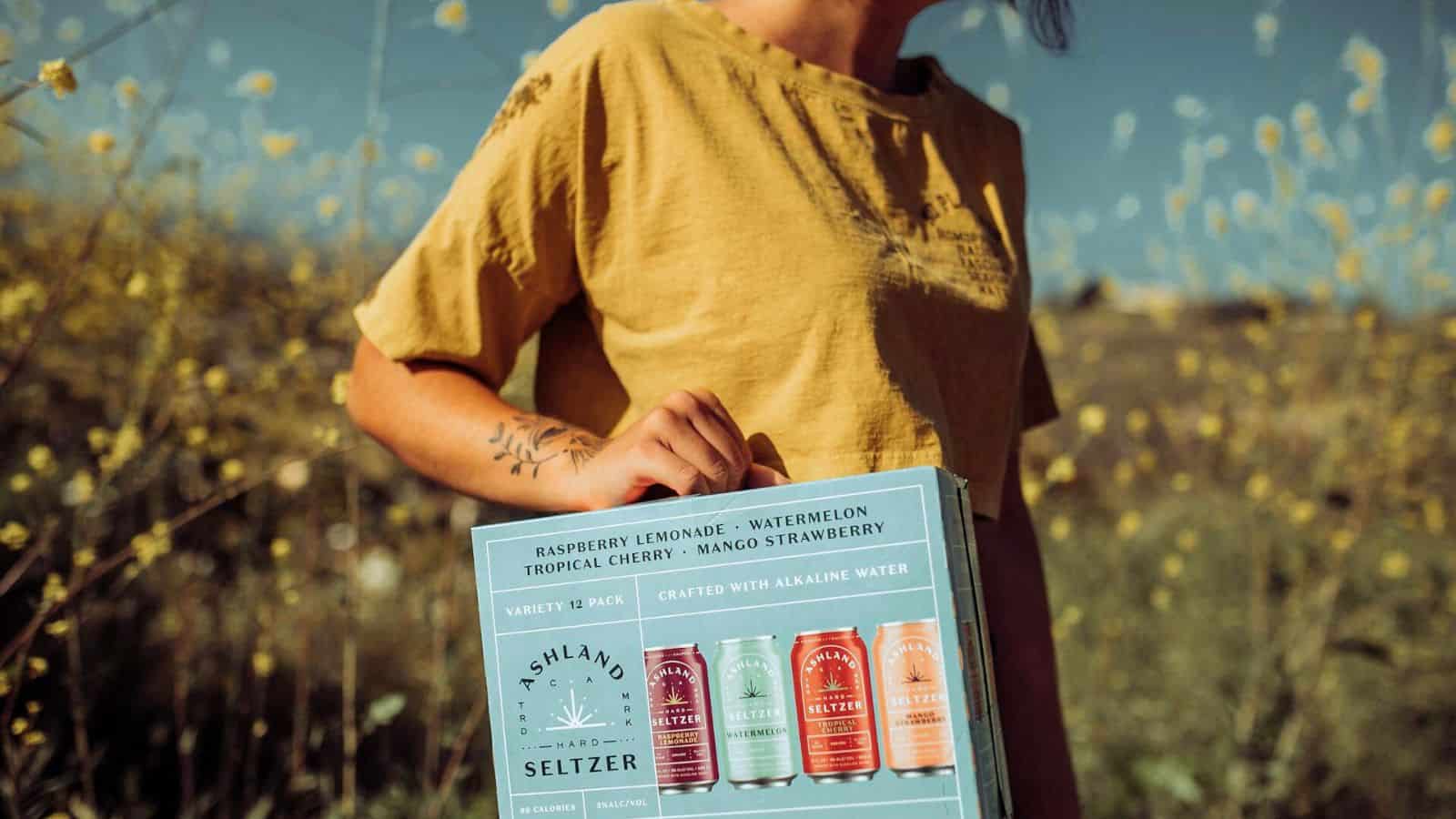 A person wearing a yellow shirt holds a variety pack of Ashland Seltzer. The pack includes flavors like Raspberry Lemonade, Tropical Cherry, Watermelon, and Mango Strawberry. In the background, a field with yellow flowers stretches under a clear blue sky.
