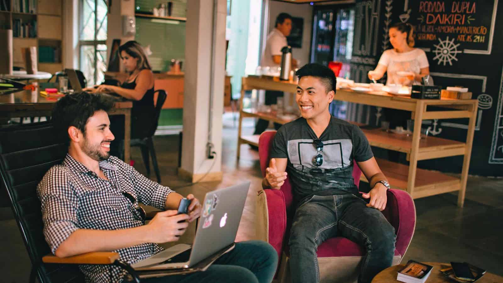 Two men are sitting on armchairs in a casual setting, engaging in conversation. One man has a laptop on his lap while the other is holding a smartphone. In the background, other people are interacting near a counter and a chalkboard wall.