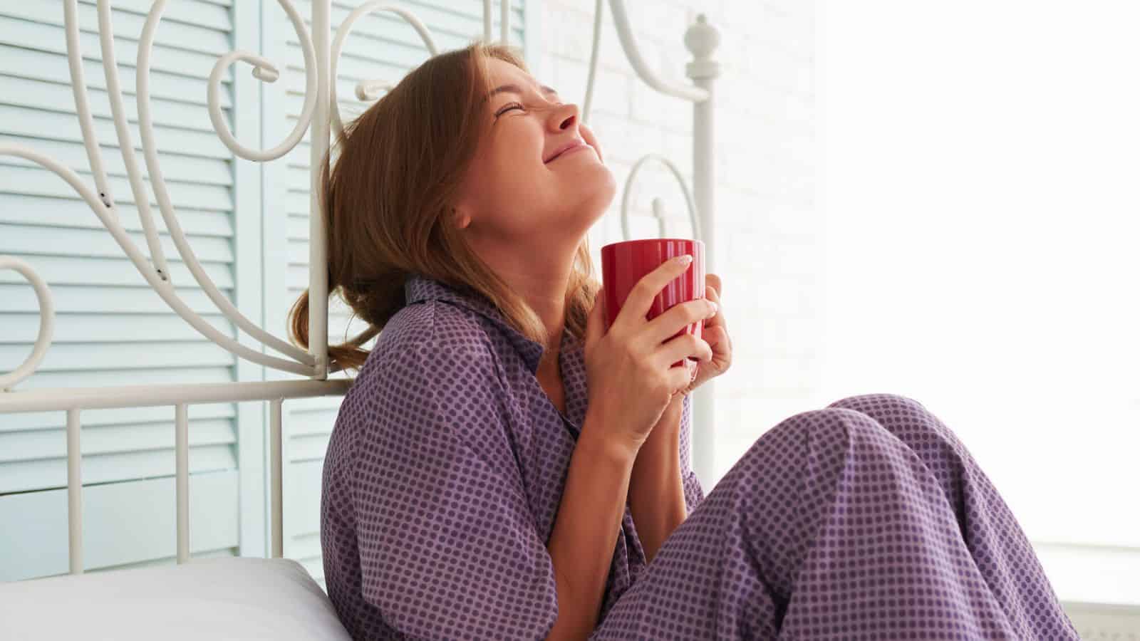 A person with brown hair sits on a bed, wearing purple pajamas and holding a red mug. They are leaning back slightly with a content expression and closed eyes. The background includes white and light blue decor.