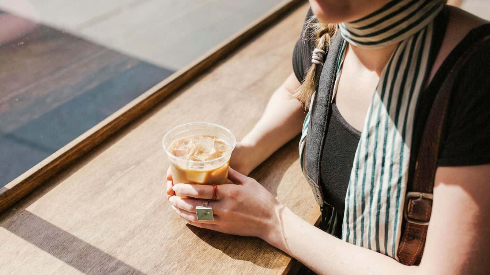 A person wearing a striped scarf and black shirt is seated at a wooden table holding a plastic cup of iced coffee with both hands. The sunlight is shining through a nearby window, casting shadows on the table.