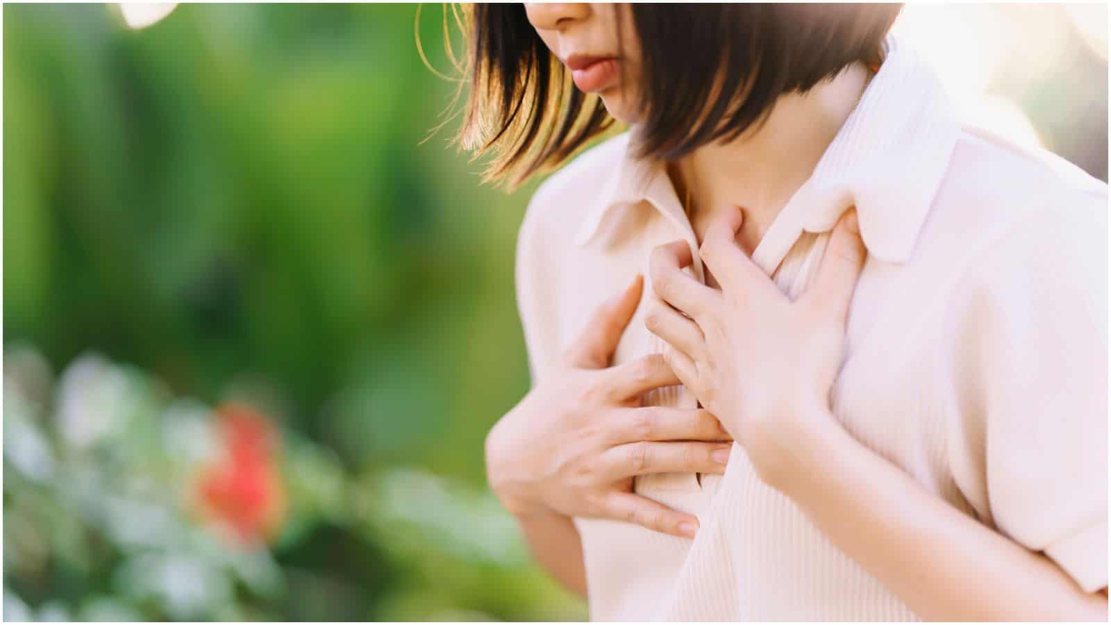 A person with shoulder-length hair holds their hands over their chest, appearing to be in discomfort. The background is blurred with greenery and flowers in soft sunlight. The person is wearing a light-colored, short-sleeve top.