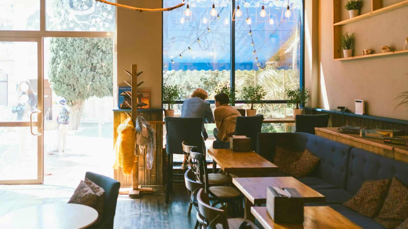 Two people sit at a high-top table by a large window in a cozy cafe. The sun shines through the windows, illuminating the wooden furniture and indoor plants. This spot charms with its mix of couches, chairs, and tables. Light bulbs hang overhead as decoration.